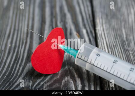 Un cuore di carta forato con una siringa medica. Sullo sfondo di tavole di pino in nero. Foto Stock