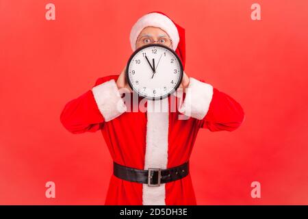 Uomo nel cappello di babbo natale che tiene l'orologio a parete vicino al viso, cinque minuti a mezzanotte, festa di nuovo anno. Studio interno isolato su sfondo rosso Foto Stock