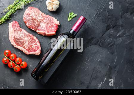 Tagli di bistecca grezzi, con bottiglia di vino rosso e bicchieri, erbe e condimenti su sfondo nero testurizzato vista laterale con spazio per il testo. Foto Stock