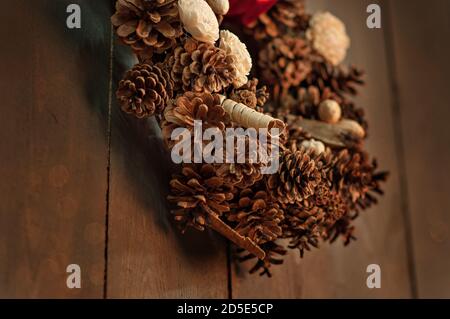Su un fondo di parete marrone di legno con bokeh sfocato in un loft scuro appende una corona fatta da mani di coni di pino, fiori secchi, elementi decorativi, Foto Stock