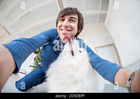 Un bel giovane in una camicia a scacchi blu ride e prende un selfie con un Husky Samoyed che leccano la guancia di un ragazzo con la sua lingua. Albero di Natale Foto Stock