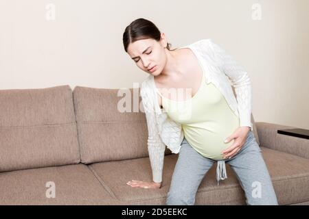 La gestante madre soffre di mal di schiena e cerca di sedersi sul divano. Foto Stock