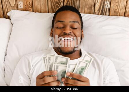 Felice uomo africano che tiene i soldi che contano i contanti che si trovano nel letto Foto Stock