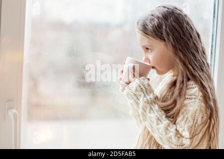 Una bambina in un pullover bianco lavorato a maglia beve cioccolata calda, cacao. Il bambino guarda fuori la finestra, attende un Natale miracolo di Capodanno, mangia Foto Stock