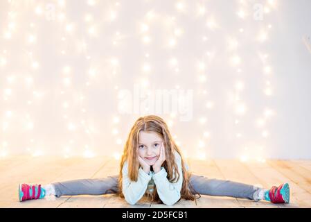 Una bella bambina bionda si siede in spago su un pavimento di legno contro uno sfondo di blurry giallo ghirlande di Natale bokeh. Carta anno nuovo. Bambino Foto Stock