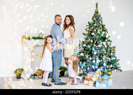 Famiglia felice, genitori abbracciando con le loro figlie vicino al camino bianco con un elegante albero di Natale in ghirlande di luci. Spazio di copia per il nuovo anno Foto Stock