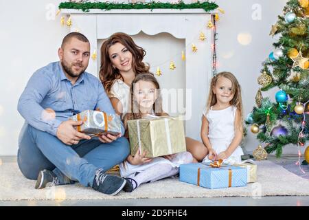 La famiglia felice sta tenendo scatole con i regali davanti ad un camino bianco con un albero di Natale elegante in ghirlande di luci. Giovane padre e beautiff Foto Stock