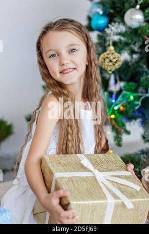 Primo piano ritratto di una bambina bionda in un vestito bianco, che tiene un regalo vicino all'albero di Natale con una ghirlanda di luci. Festa di Capodanno entro Foto Stock