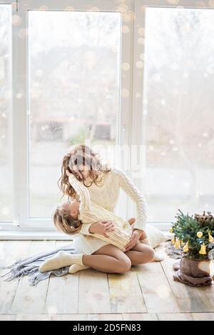 Una bella madre sta giocando con la figlia felice alla finestra panoramica nel maglione lavorato a maglia vicino all'albero di Natale. Una giovane donna abbraccia la ragazza Foto Stock
