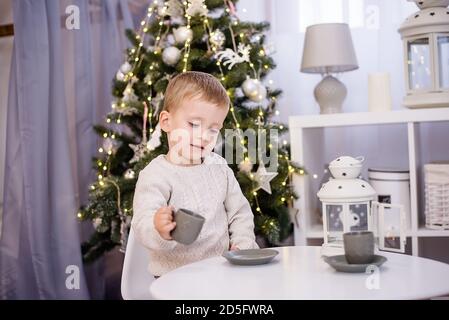 Un ragazzino in un pullover bianco gioca il tè, beve il cacao da una tazza grigia al suo tavolo bianco nella stanza dei bambini. Sullo sfondo è un Natale Foto Stock