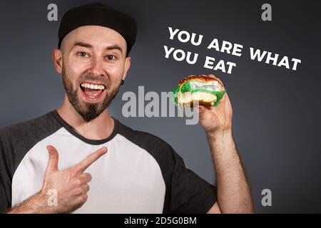 Il concetto di fast food ed ecologia. Un uomo in una cuffia e una barba indica felicemente un hamburger nella sua altra mano. Sfondo nero. TESTO SEI CIÒ CHE Foto Stock