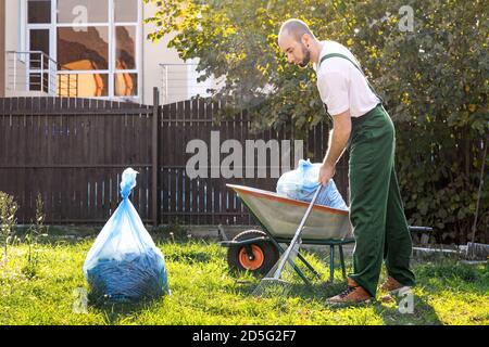 Il giardiniere nell'uniforme verde sta pulendo il yard.on l'erba è un carrello con composto e un pacchetto di immondizia. Foto Stock