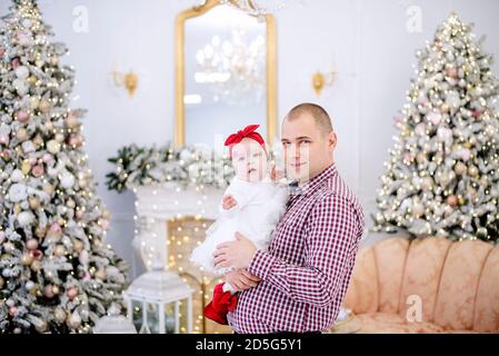 Un giovane tiene un bambino tra le braccia. Padre gioca con una ragazza in un vestito bianco, la abbraccia, bacia sullo sfondo di Natale alberi festosi Foto Stock
