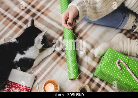 Giovane donna si siede su plaid in maglia accogliente pullover bianco lana, calzini e avvolge regalo di Natale in polka dot wrapping carta. Divertente tuxe bianco e nero Foto Stock