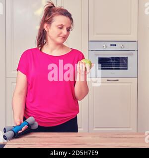 Ragazza dopo un allenamento serale in cucina con una mela in mano. Il concetto di dieta e di allenamento sportivo a casa in quarantena durante il coron Foto Stock