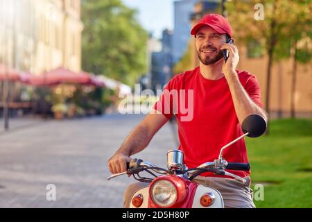 Giovane allegro uomo caucasico di consegna che parla al telefono mentre si siede su scooter sulla strada soleggiata. Concetto di consegna degli alimenti Foto Stock