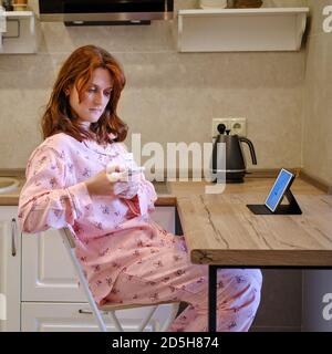 Una giovane donna in pigiama lavora online da casa. La ragazza seduta al tavolo in cucina chiama al telefono. Lavoro da casa durante la quarantena e. Foto Stock