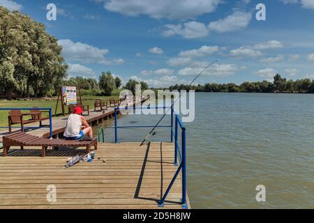 VILKOVE, UCRAINA - 05 AGOSTO 2020: L'uomo pesca sulla riva del Danubio in banchina di legno con posti confortevoli per il riposo e la pesca in un turista r Foto Stock