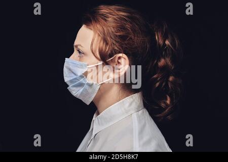 Donna in una maschera protettiva su sfondo scuro, ritratto di profilo. Primo piano di una ragazza dai capelli rossi in una camicia bianca Foto Stock