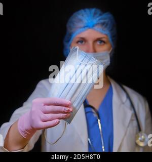 Medico con una maschera medica in mano, primo piano. Nurse dà maschere facciali su uno sfondo nero. Concetto di abbigliamento protettivo pandemico coronavirus Foto Stock