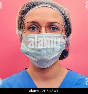 Infermiere in uniforme blu e maschera protettiva su sfondo rosso. Ritratto di una dottoressa in occhiali e un cappello medico, closeup.. Foto Stock