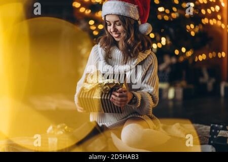 Natale, Capodanno. Bella donna in caldo maglione, calze e cappello di natale, seduta sul pavimento a casa con scatole regalo e albero di nuovo anno e luci Foto Stock