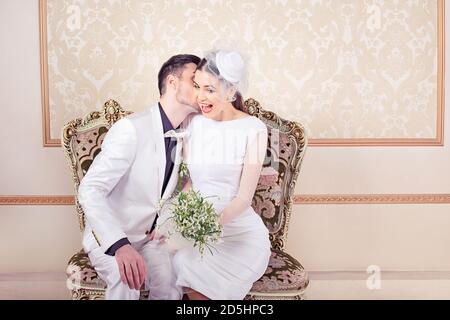 Moderno sposo di classe che dice un segreto al suo velo sposa e vestito con bouquet floreale guardando eccitato da quello che lei sta sentendo seduto sul divano in un Foto Stock