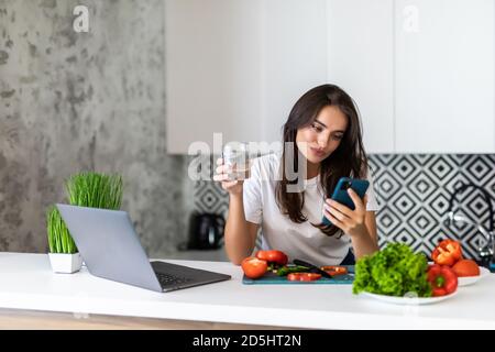 Ritratto di giovane donna che cucina verdure fresche tenendo per telefono per l'ordinazione di prodotti biologici agricoli in cucina Foto Stock
