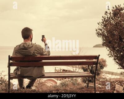 Uomo con retro guardando lo schermo del telefono mentre si siede su una panchina all'aperto, nel parco vicino al mare Foto Stock