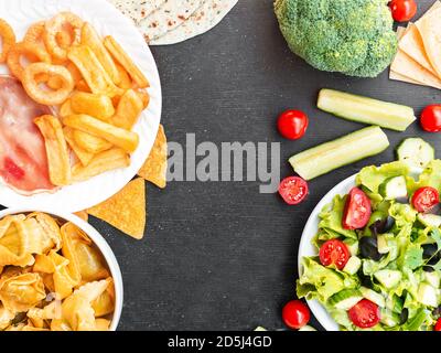 Cibo sano contro malsano e concetto di dieta. Cibo spazzatura e verdure insalata verde in ciotole isolato su sfondo nero studio con spazio di copia. Piatto Foto Stock