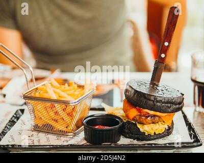 Cibo malsano e concetto di fast food. Cibo spazzatura, hamburger nero e patate fritte francesi di fronte alla silhouette uomo su un tavolo in un ristorante. Visualizza Foto Stock