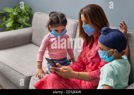 UN FIGLIO SIKH, MADRE E FIGLIA INDOSSANO MASCHERE E SI SIEDE SUL DIVANO MENTRE SI GUARDA IL TELEFONO CELLULARE Foto Stock