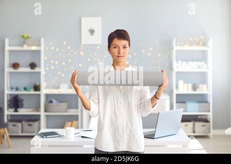 Giovane donna d'affari che tiene il tappeto di yoga in mani in piedi in un ambiente accogliente ufficio e guardando la fotocamera Foto Stock