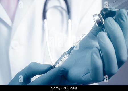 Vista ravvicinata della siringa e della fiala di vetro a mani vestite con guanti di gomma blu, medico vestito con accappatoio bianco su sfondo sfocato Foto Stock