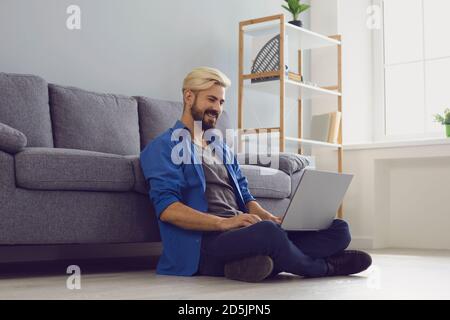Un uomo sorridente seduto a casa sul pavimento con un computer portatile e. lavorare online Foto Stock