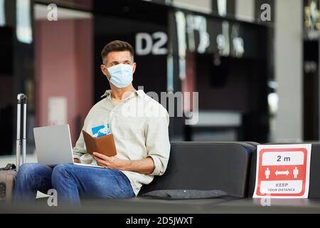 Giovane uomo in maschera protettiva con bagaglio e biglietti seduti in aeroporto durante la pandemia Foto Stock