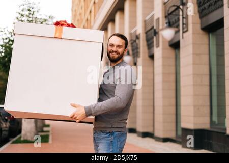 Ampio ritratto di allegro delivery uomo che tiene grande festa scatola con arco rosso all'aperto Foto Stock