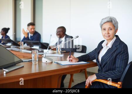 Ritratto di uomo d'affari maturo che guarda la macchina fotografica mentre si siede a. conferenza con i suoi colleghi sullo sfondo Foto Stock