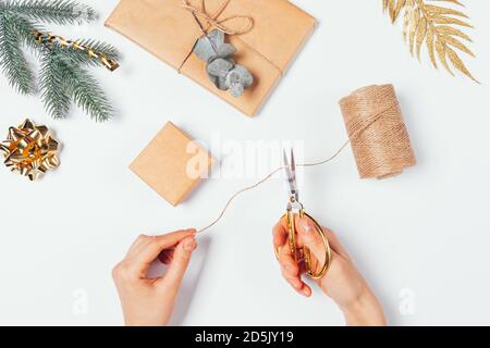 Le mani femminili tengono le forbici tagliando lo spago per fare l'imballaggio fatto in casa per i regali di Natale vicino alle scatole del mestiere marrone, le decorazioni dorate, l'abete e l'euca Foto Stock