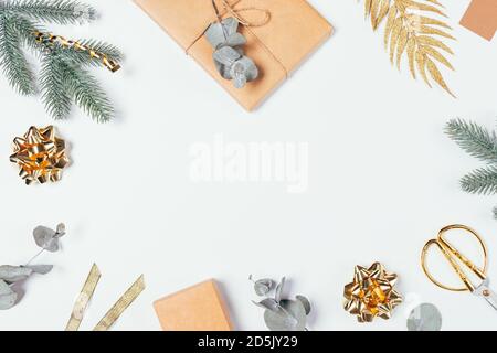 Cornice piatta con regali di Natale in carta marrone artigianale, rami di abete verde e eucalipto, nastri ornamenti dorati e forbici su erba bianca Foto Stock