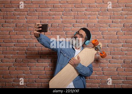 Un hipster prende un selfie sul suo smartphone con la longboard su uno sfondo di muro di mattoni. Foto Stock