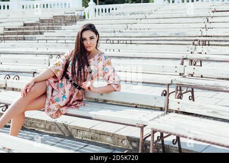 Bella donna brunette in un abito con fiori si siede su una panca da sola Foto Stock