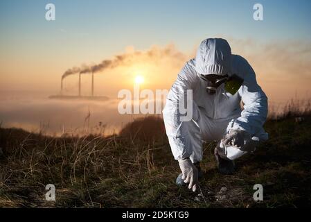 Ambientalista accovacciato che lavora all'aperto sulla collina raccogliendo campioni per il suo studio, i tubi fumanti di una centrale termica sono sullo sfondo, concetto di emissioni industriali Foto Stock