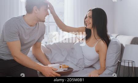 Shot di un giovane uomo premuroso che porta vassoio con colazione a letto alla sua bella partner femminile. Ideale relazione tenera e caffè e croissant in Foto Stock