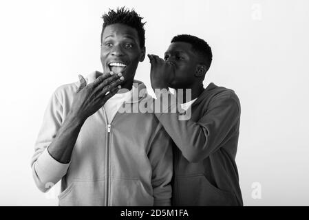 Giovane uomo africano sussurrando a felice uomo africano sorridente mentre pensare e guardare scioccato Foto Stock