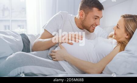 Felice giovane coppia coccolandosi insieme nel letto, giovane donna è incinta, partner amorevole si appoggia e ascolta un bambino che si muove ed è Heartbeat, Kisses Foto Stock