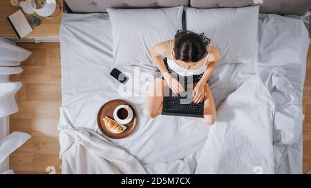 Efficient Young Millennial Girl seduta su un letto al mattino, utilizza il computer portatile e mangia croissant e bevande caffè per la prima colazione. Dall'alto verso il basso Foto Stock