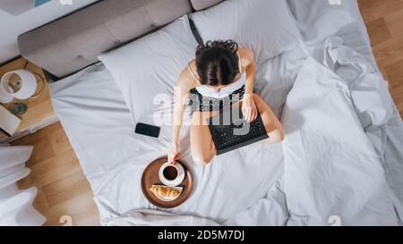 Efficient Young Millennial Girl seduta su un letto al mattino, utilizza il computer portatile e mangia croissant e bevande caffè per la prima colazione. Dall'alto verso il basso Foto Stock