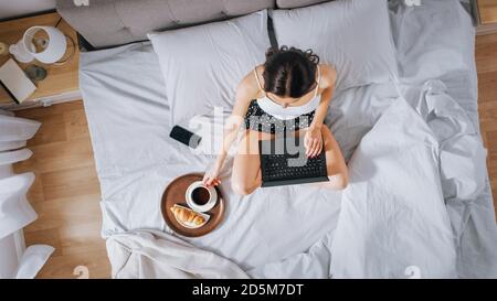 Efficient Young Millennial Girl seduta su un letto al mattino, utilizza il computer portatile e mangia croissant e bevande caffè per la prima colazione. Dall'alto verso il basso Foto Stock