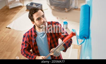 L'uomo in jeans marroni e camicia rossa controllata sta dipingendo una parete con un rullo. Il colore della vernice è azzurro. Ristrutturazione delle camere in casa. Ripresa dall'alto. Foto Stock